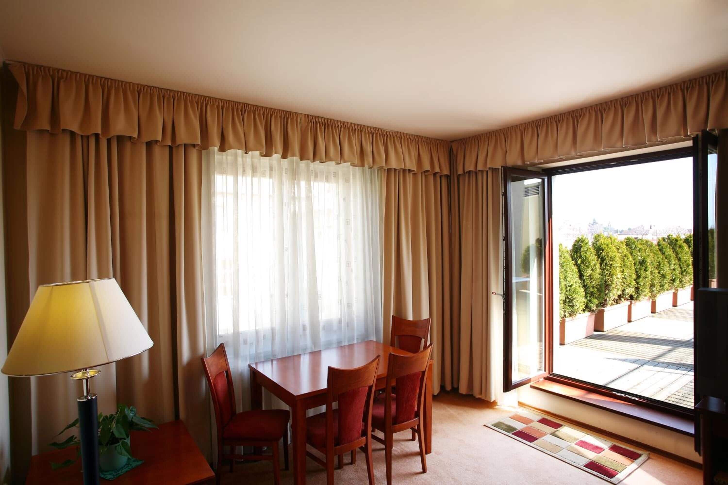 Ein Hotelzimmer mit Esstisch, vier Stühlen, einer Lampe und Blick auf einen Garten durch ein großes Fenster.