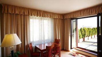 Ein Hotelzimmer mit Esstisch, vier Stühlen, einer Lampe und Blick auf einen Garten durch ein großes Fenster.