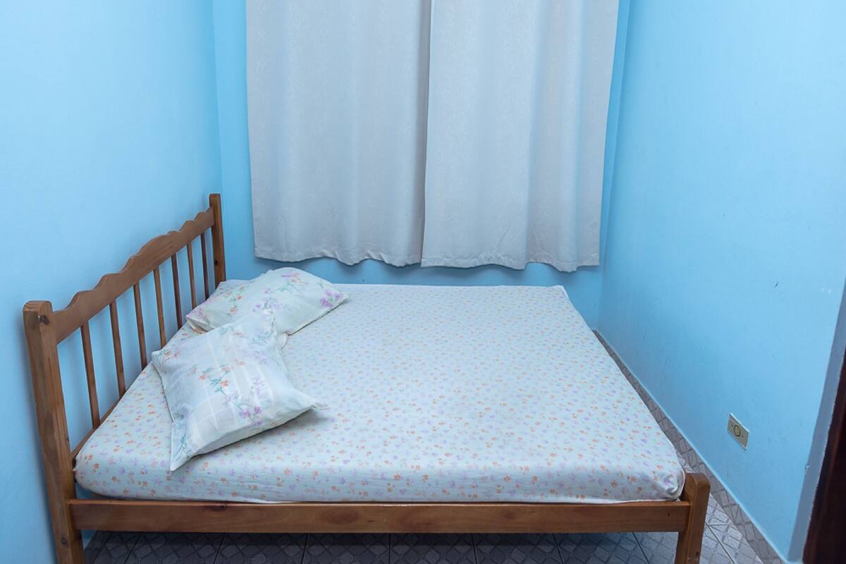 Ein Einzelbett mit Holzrahmen und einer Matratze mit Blumenmuster, vor einer blauen Wand mit einem weißen Vorhang.