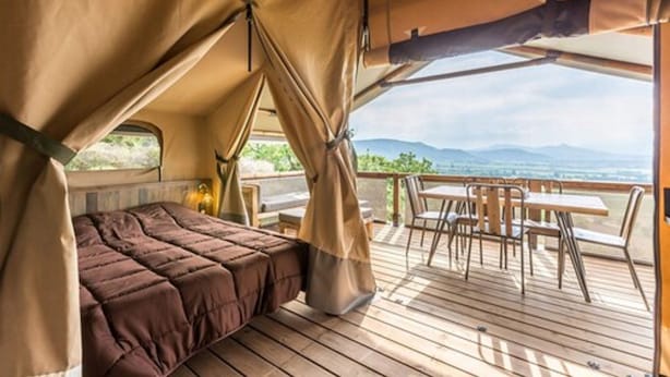Un alloggio in tenda con un letto, un tavolo e sedie su un pontile di legno, affacciato su un paesaggio montuoso.