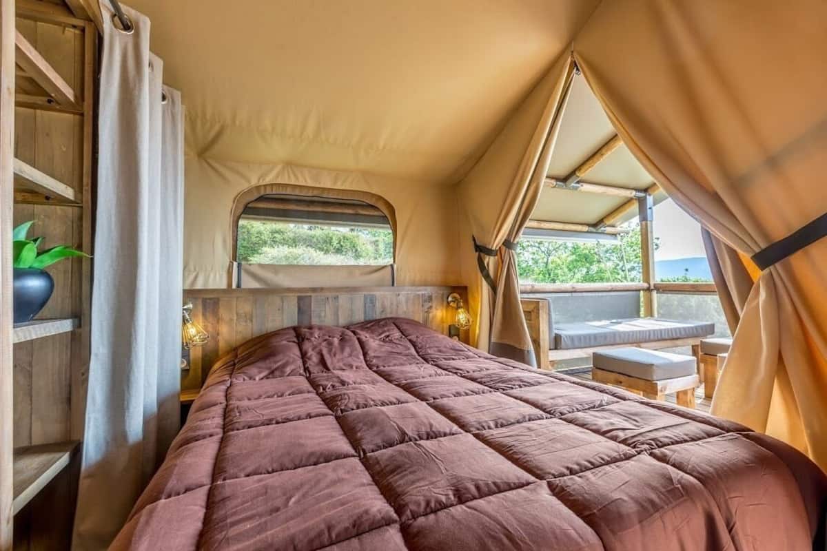 Une pièce accueillante aménagée comme une tente, avec un grand lit, des étagères en bois, une petite table et une fenêtre donnant sur un paysage verdoyant.