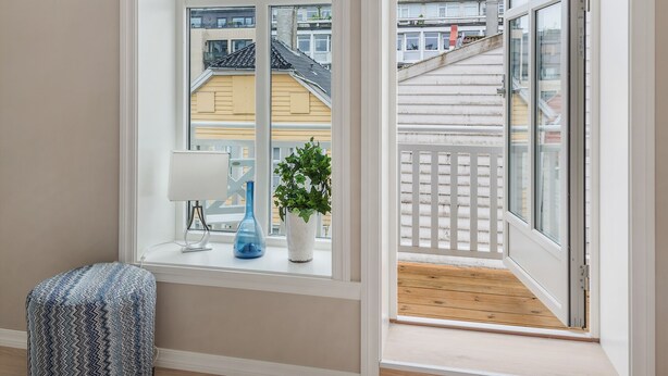A room with a large window, a small table with a lamp and a plant, and a patterned ottoman. - Fosswinckel Apartments (Bergen)