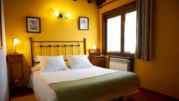A bed with white linens and a green throw, a nightstand with a lamp, and a window with curtains. - Posada La Vieja Escuela (San Vicente de la Barquera)