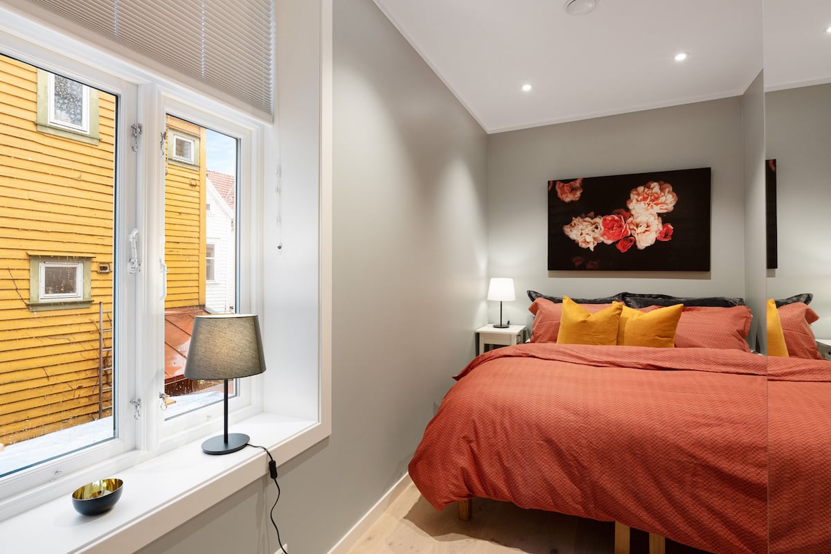 Un dormitorio con un ventanal grande, una cama con ropa de cama naranja, una mesita de noche con una lámpara y un cuadro floral en la pared.
