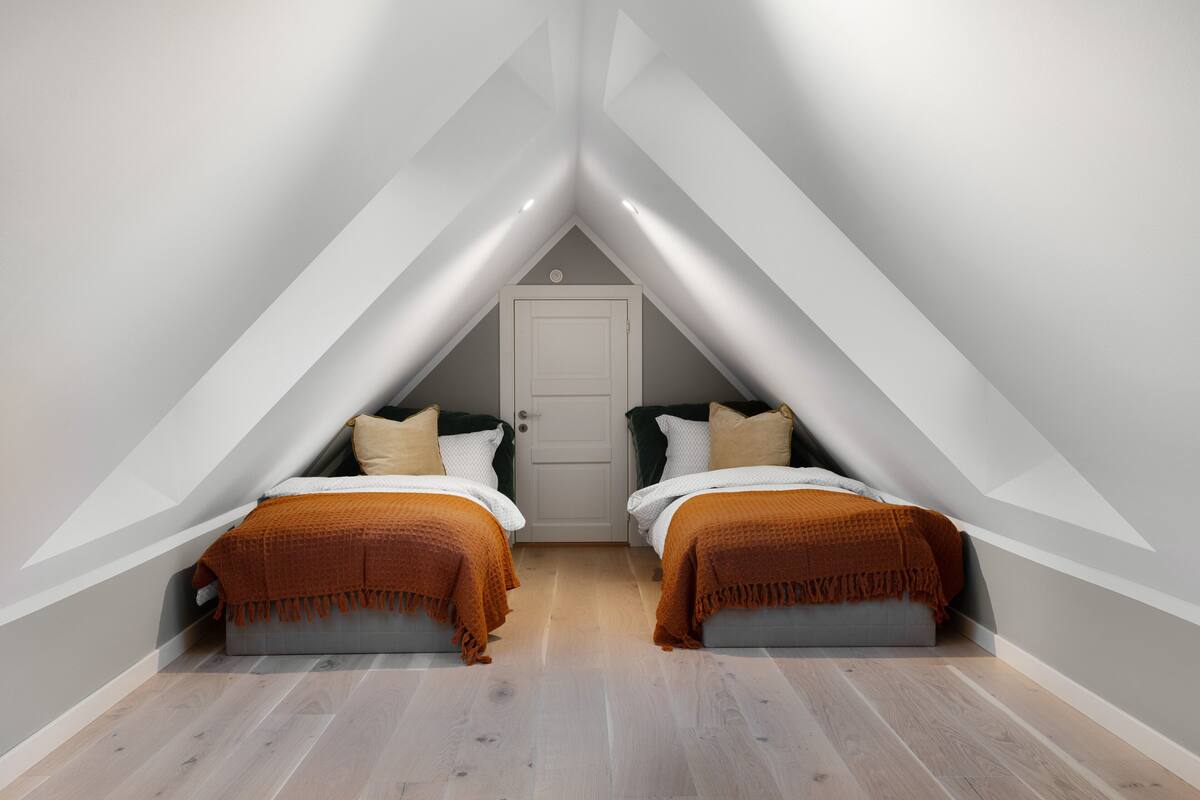 Un pequeño dormitorio en ático, con forma de triángulo, que tiene dos camas, una puerta y suelo de madera.