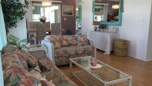 A living room with a sofa, a glass coffee table, and a wicker chair. There is a mirror on the wall and a plant in the corner. - Walk to the Beach at Nettles Island 4 bed/2 bath sleeps 8 (Jensen Beach)