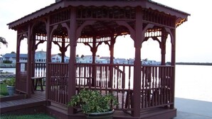 A gazebo with a roof and railings, situated on a grassy area with a view of a body of water and buildings in the background. - Walk to the Beach at Nettles Island 4 bed/2 bath sleeps 8 (Jensen Beach)