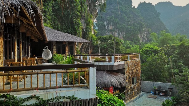 A rustic wooden building with a thatched roof, surrounded by lush greenery and steep cliffs. - Trang An Ecolodge (Tay Hoa Lu)