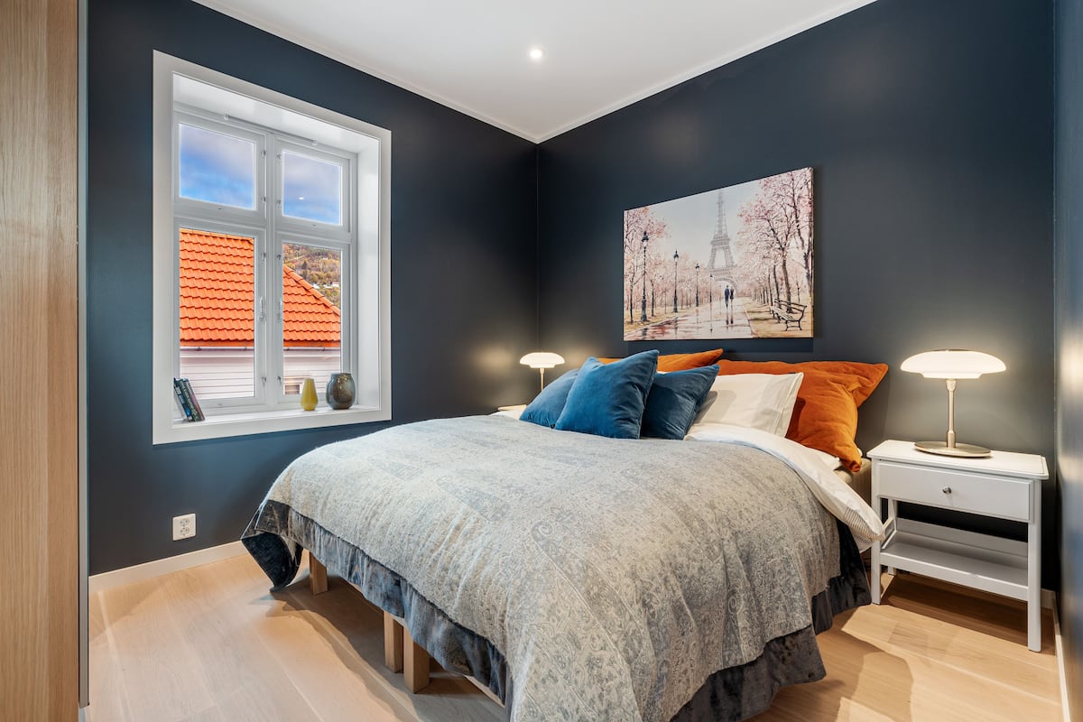 A bedroom with a large bed, a nightstand with a lamp, a window with a view of a red-roofed building, and a painting of a park scene.