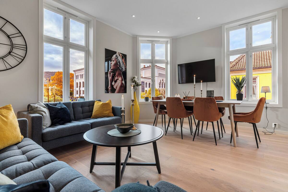 A modern living room with a sofa, dining table, and chairs, large windows with a city view, and a flat-screen TV mounted on the wall.
