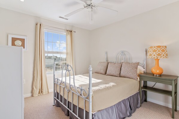 Une chambre à coucher comprenant un lit, une table de chevet, une lampe, un ventilateur de plafond et une fenêtre avec des rideaux.