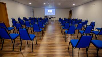 A conference room with rows of blue chairs, a wooden floor, and a screen displaying 'tri Hotel Chapecó'.