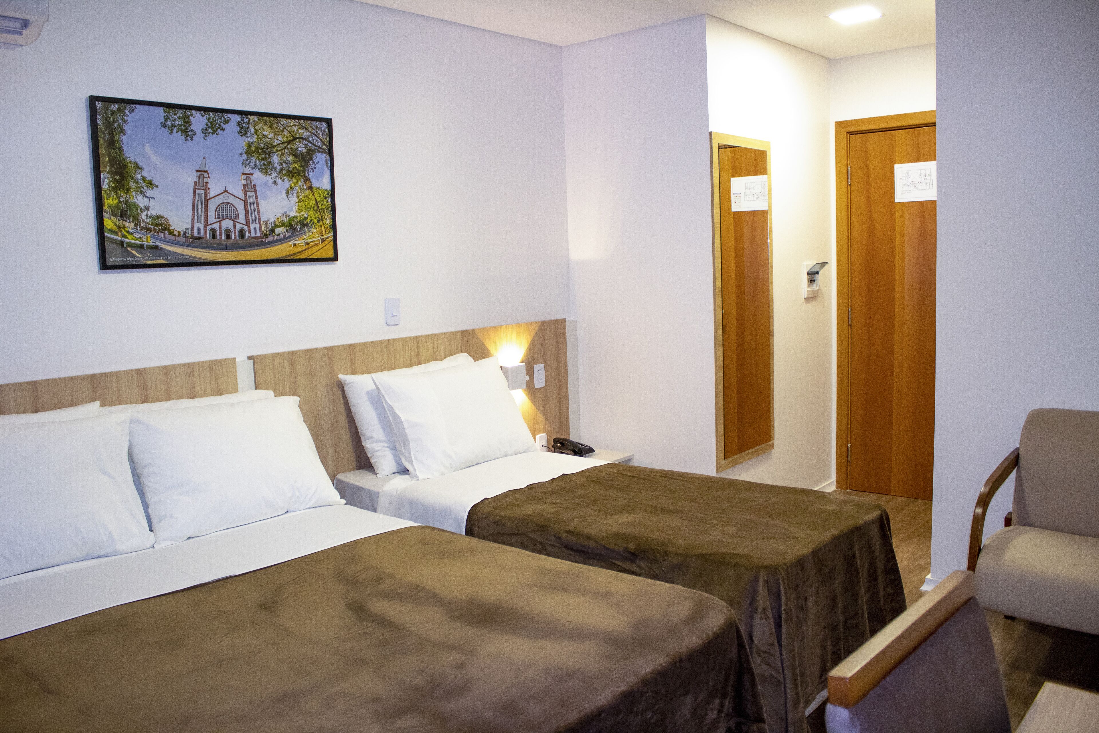 Una habitación de hotel con dos camas, un cabecero de madera, una mesita de noche, una silla y un cuadro de un edificio en la pared.