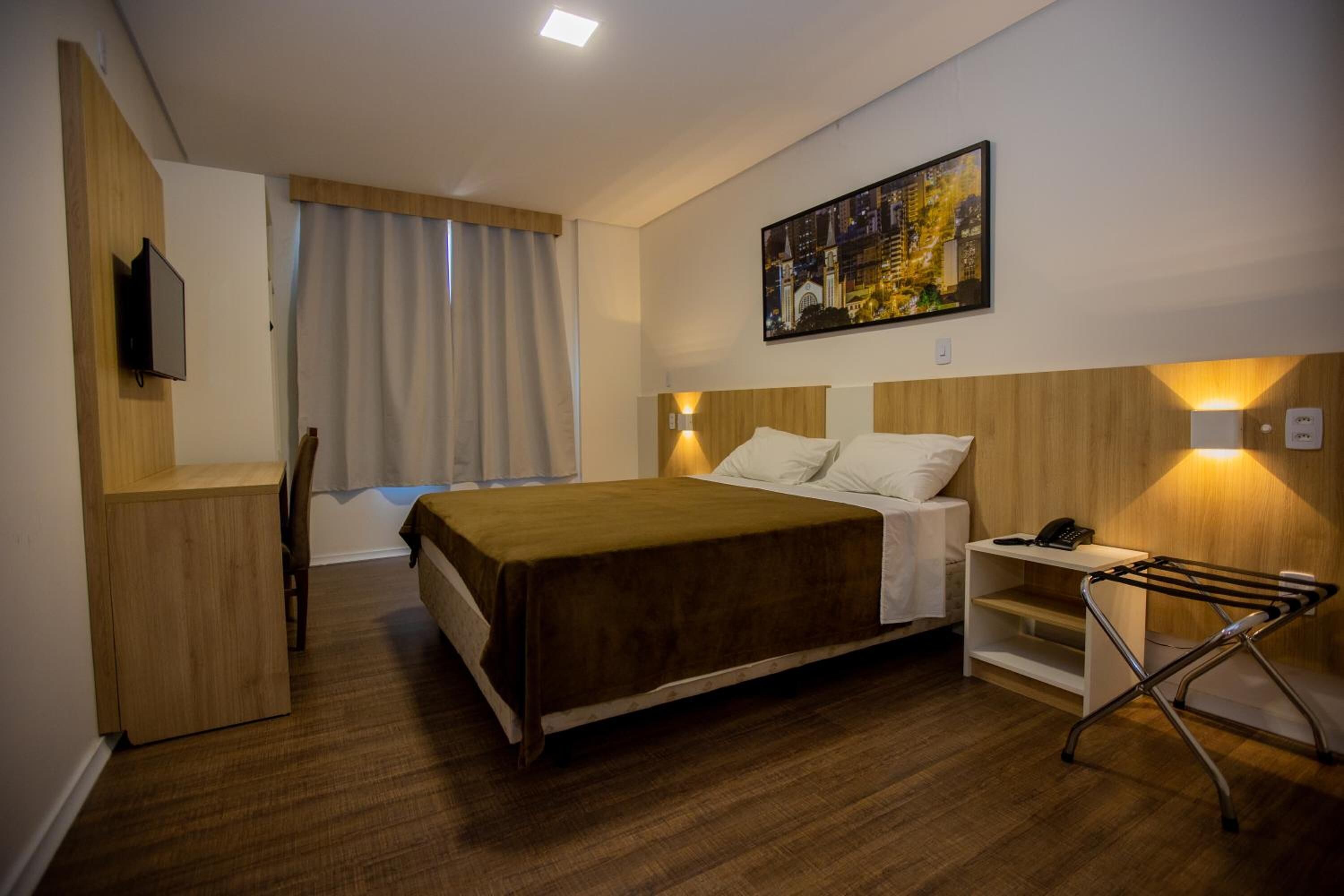 A hotel room with a bed, a desk, a chair, a TV, and a painting on the wall.
