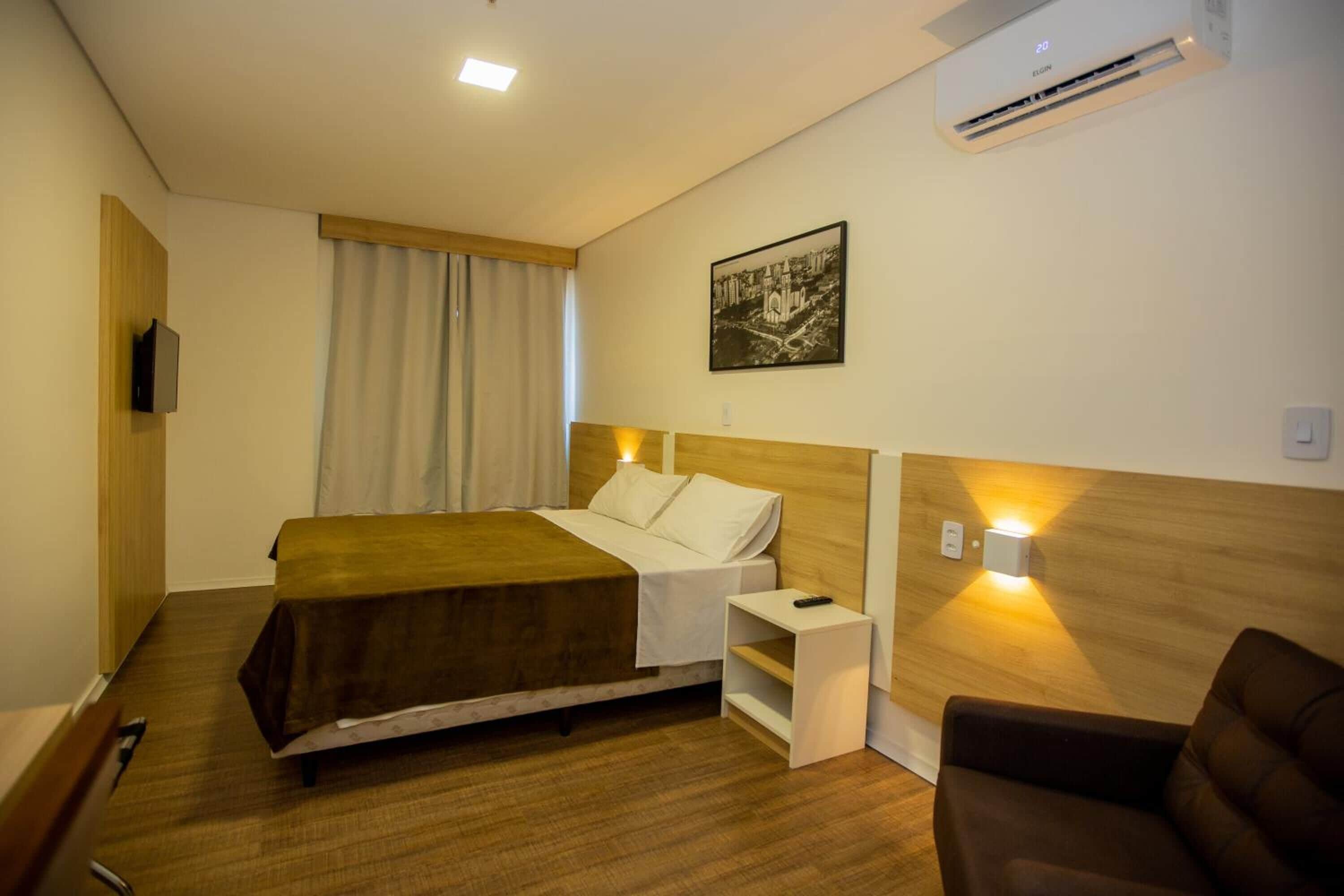 A hotel room with a bed, bedside table, lamp, and a wall-mounted air conditioner.