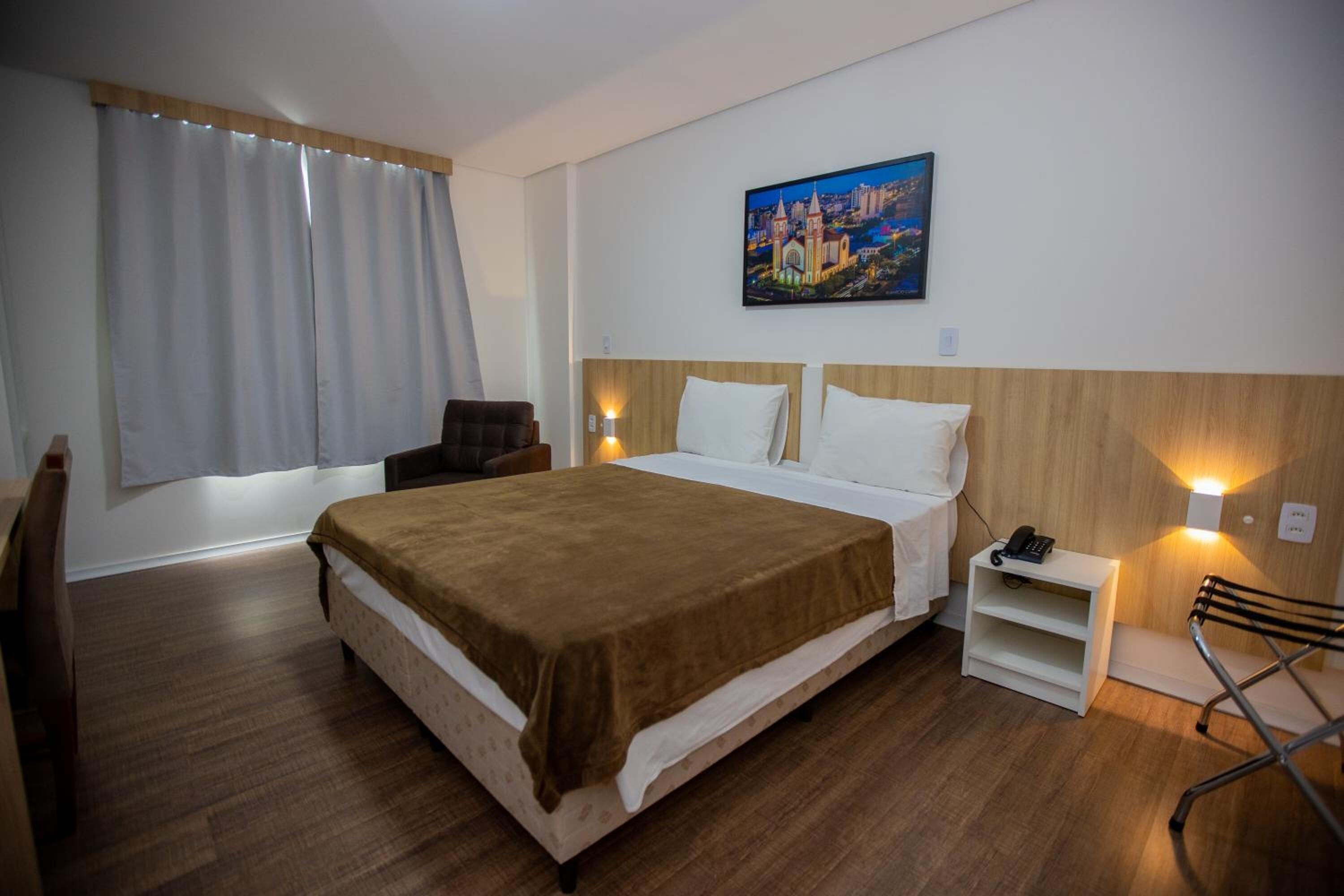 Una habitación de hotel con una cama grande, una silla, una mesita de noche con un teléfono y un cuadro en la pared.