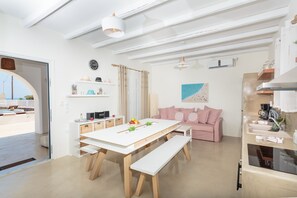 A modern kitchen with white cabinetry, a dining table with benches, a pink sofa, and a view through an open door to a balcony.