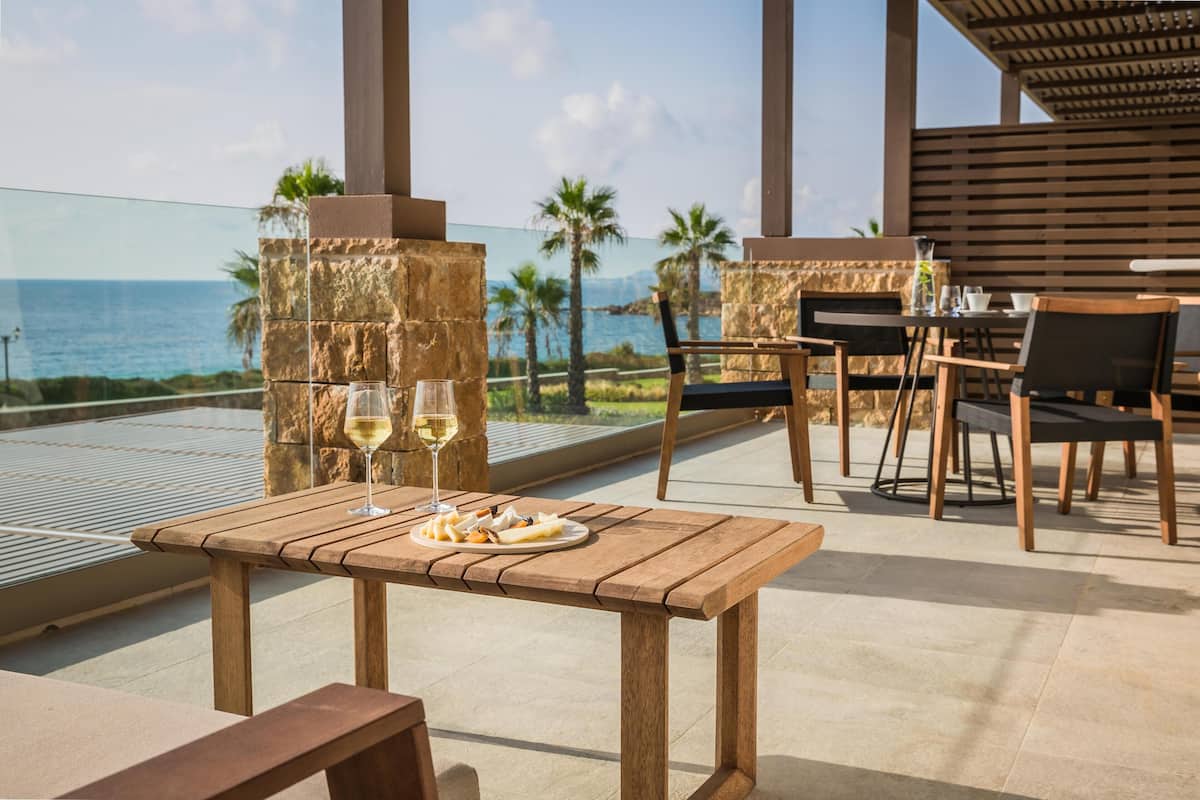 Een houten tafel met een bord eten en twee glazen wijn, voor een achtergrond van palmbomen en de zee.