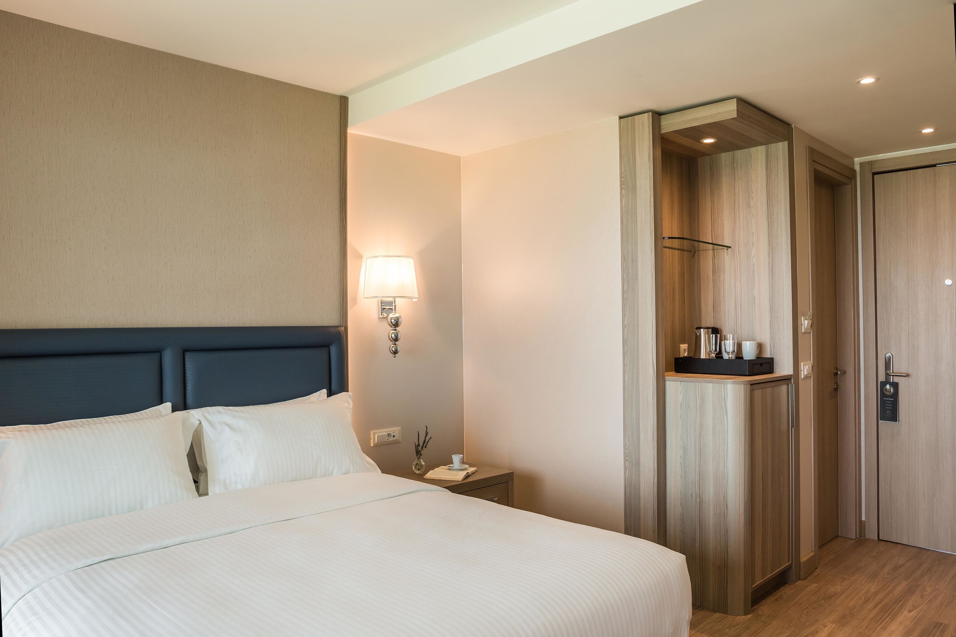 A hotel room with a large bed, a bedside table, a lamp, a wardrobe, and a small shelf with items.