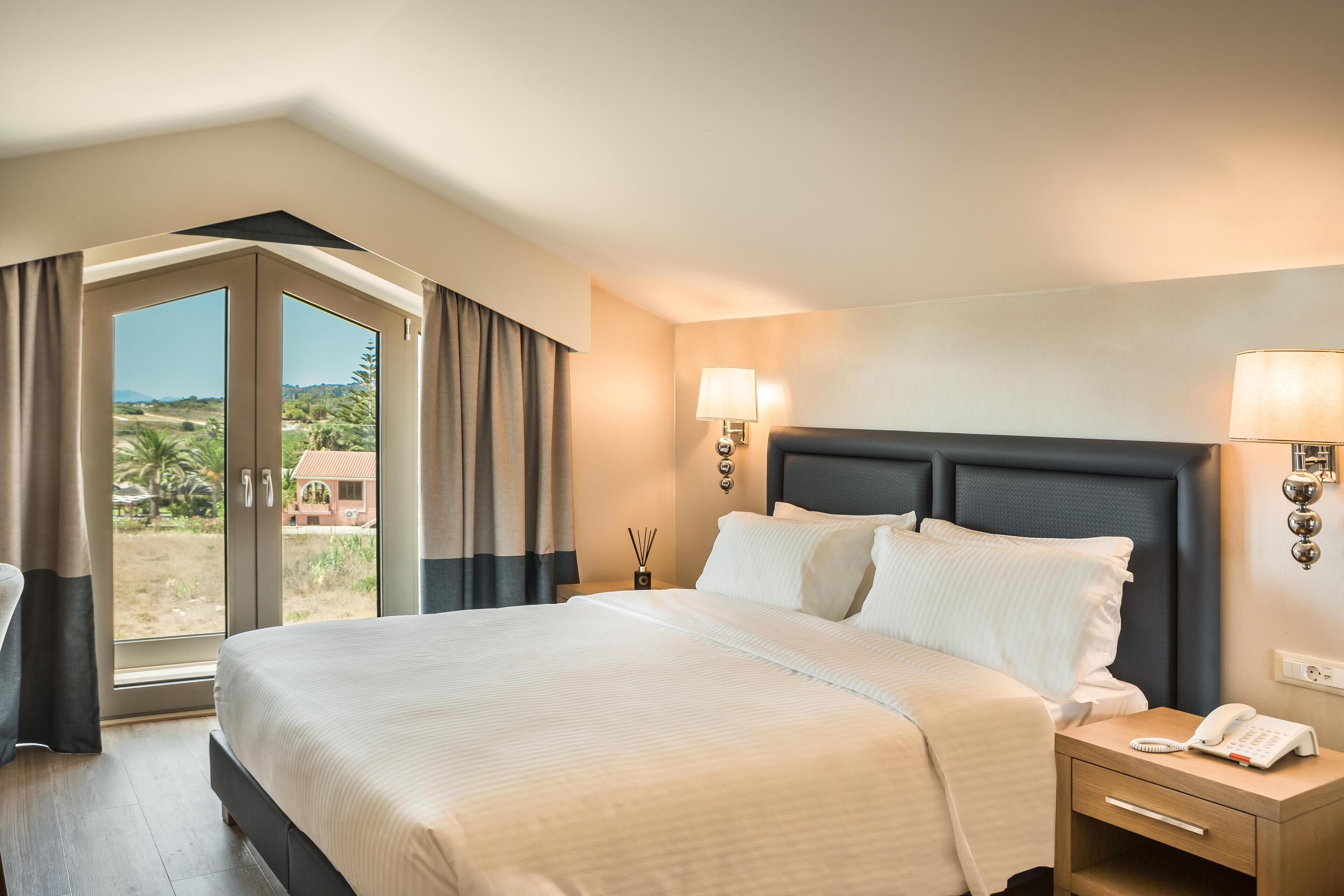 A hotel room with a large bed, a bedside table with a lamp, and a view of the outdoors.