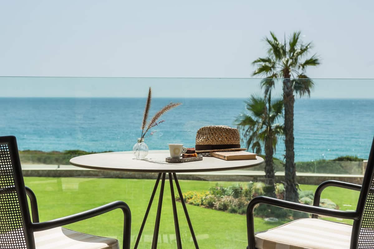 Een ronde tafel met een vaas, een kopje en een mandje op een balkon met uitzicht op de oceaan.