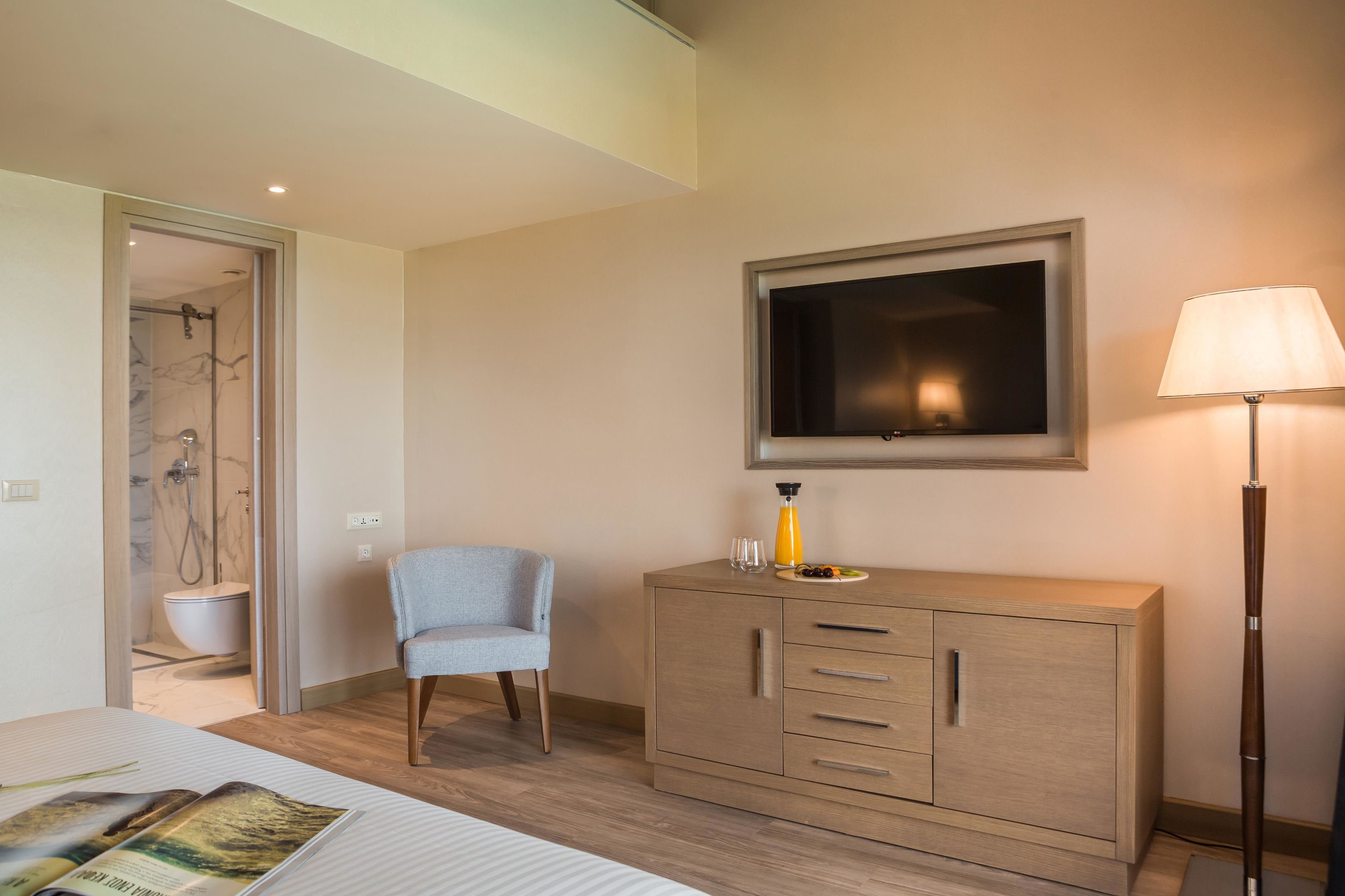A hotel room with a bed, a wooden cabinet, a chair, a television, a lamp, and a shower area.