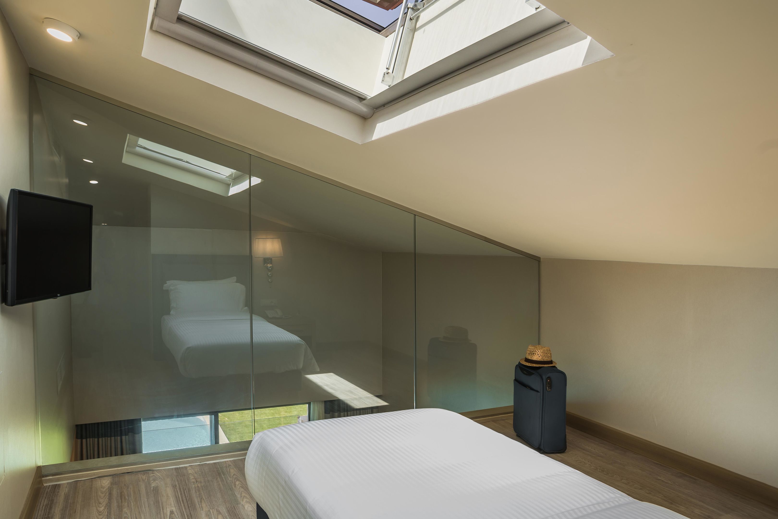 A modern hotel room with a large bed, a flat-screen TV, a glass partition, and a skylight.