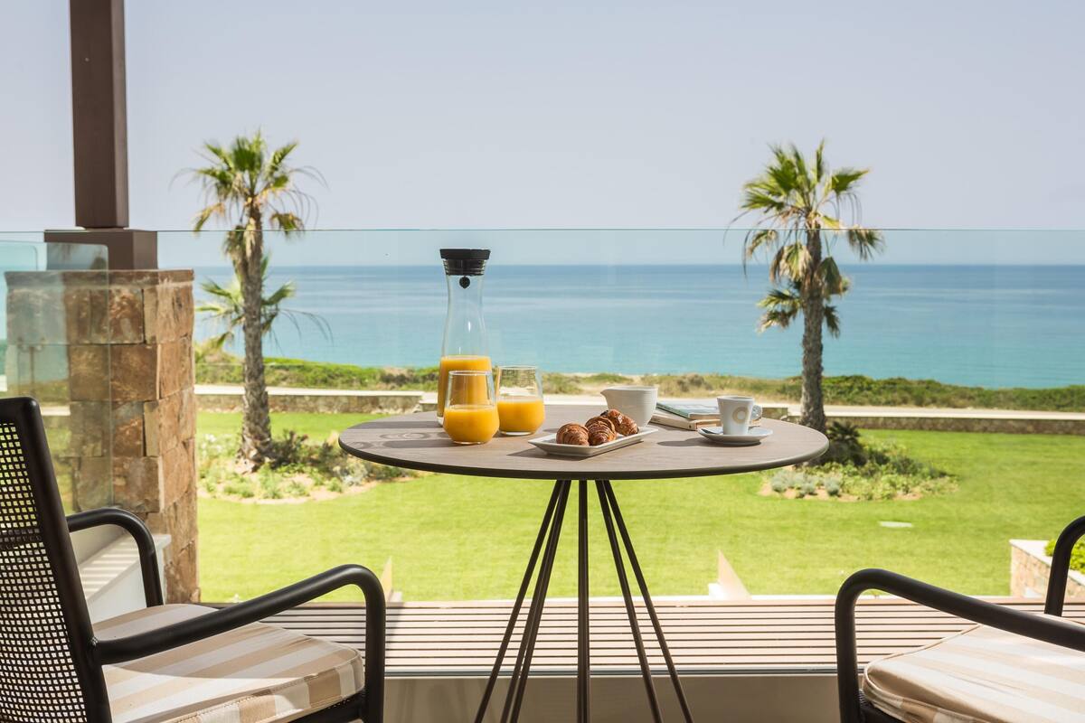 Een tafel met ontbijtartikelen en een glas sinaasappelsap op een balkon met uitzicht op de oceaan.