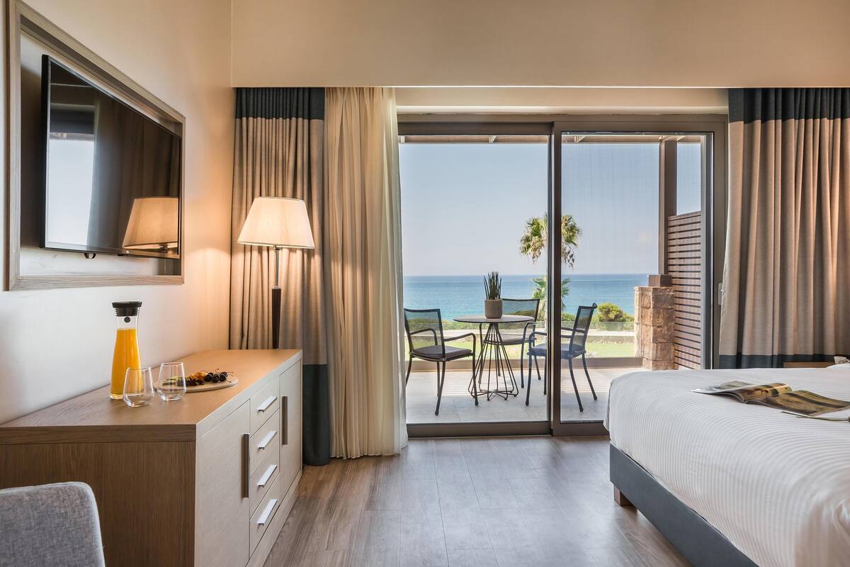 Een hotelkamer met een bed, een bureau met lamp, een televisie en uitzicht op zee.