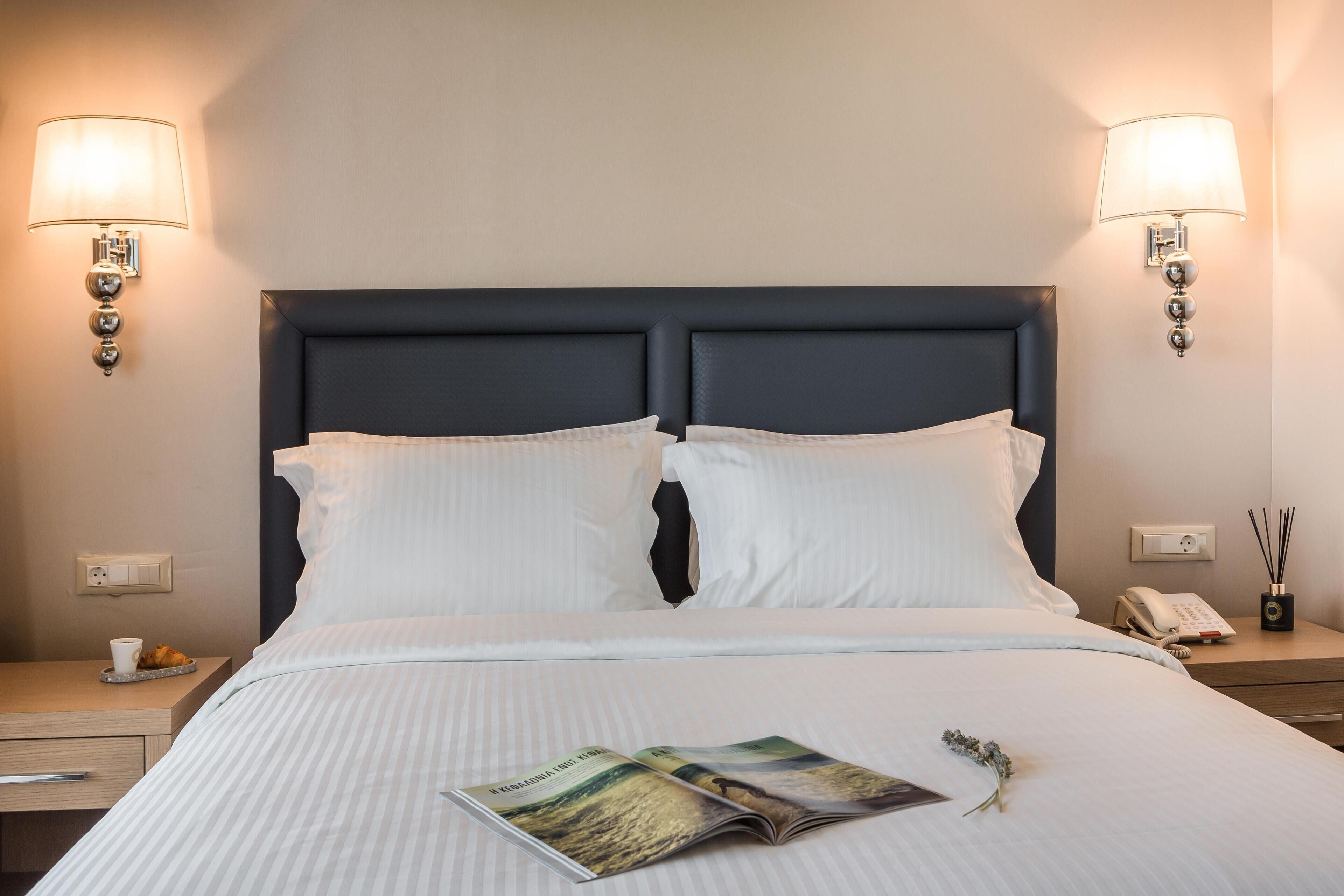 A neatly made bed with white linens, two wall-mounted lamps, a bedside table with a phone and a small vase, and a magazine on the bed.