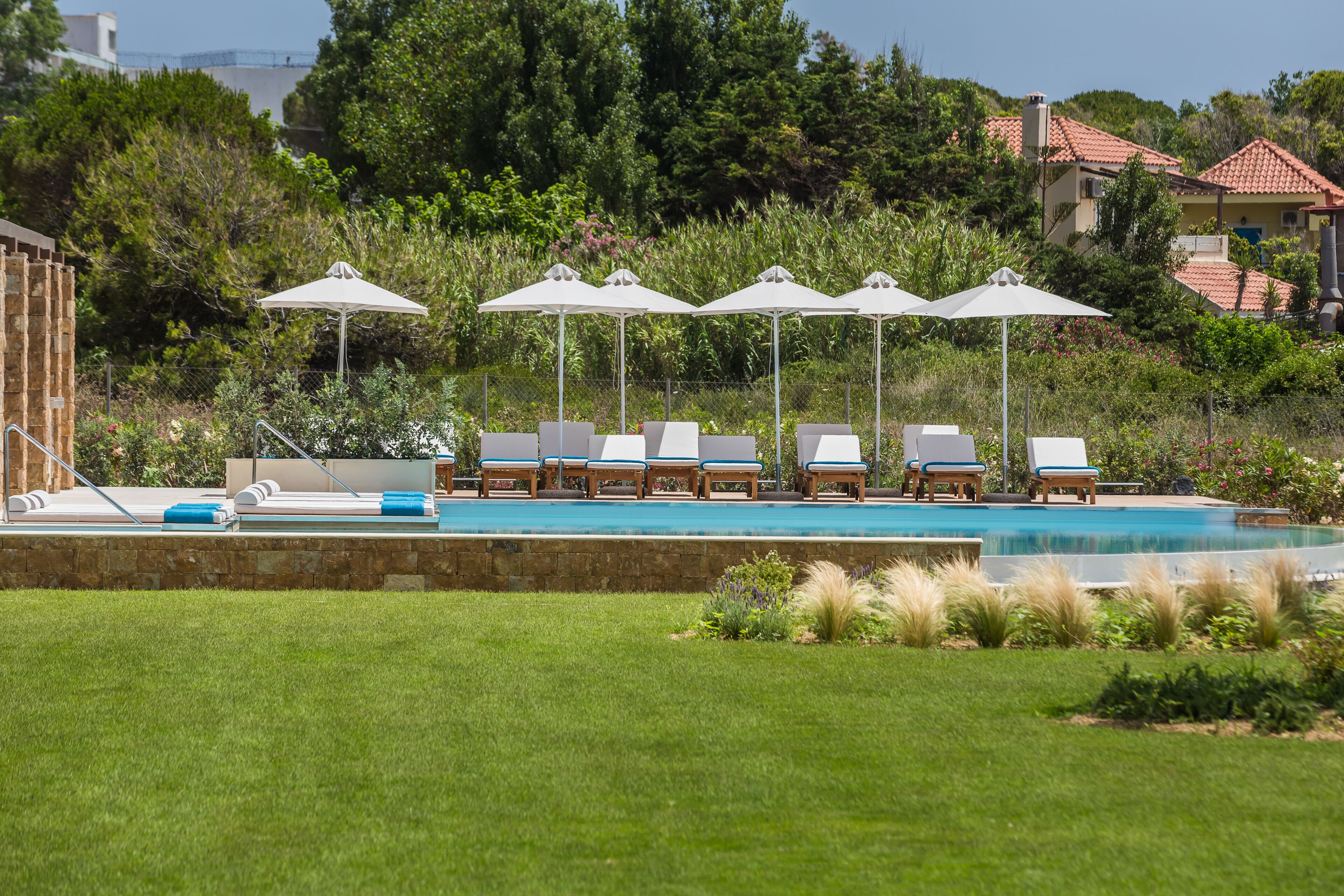 A pool area with lounge chairs and umbrellas, surrounded by greenery and buildings in the background.