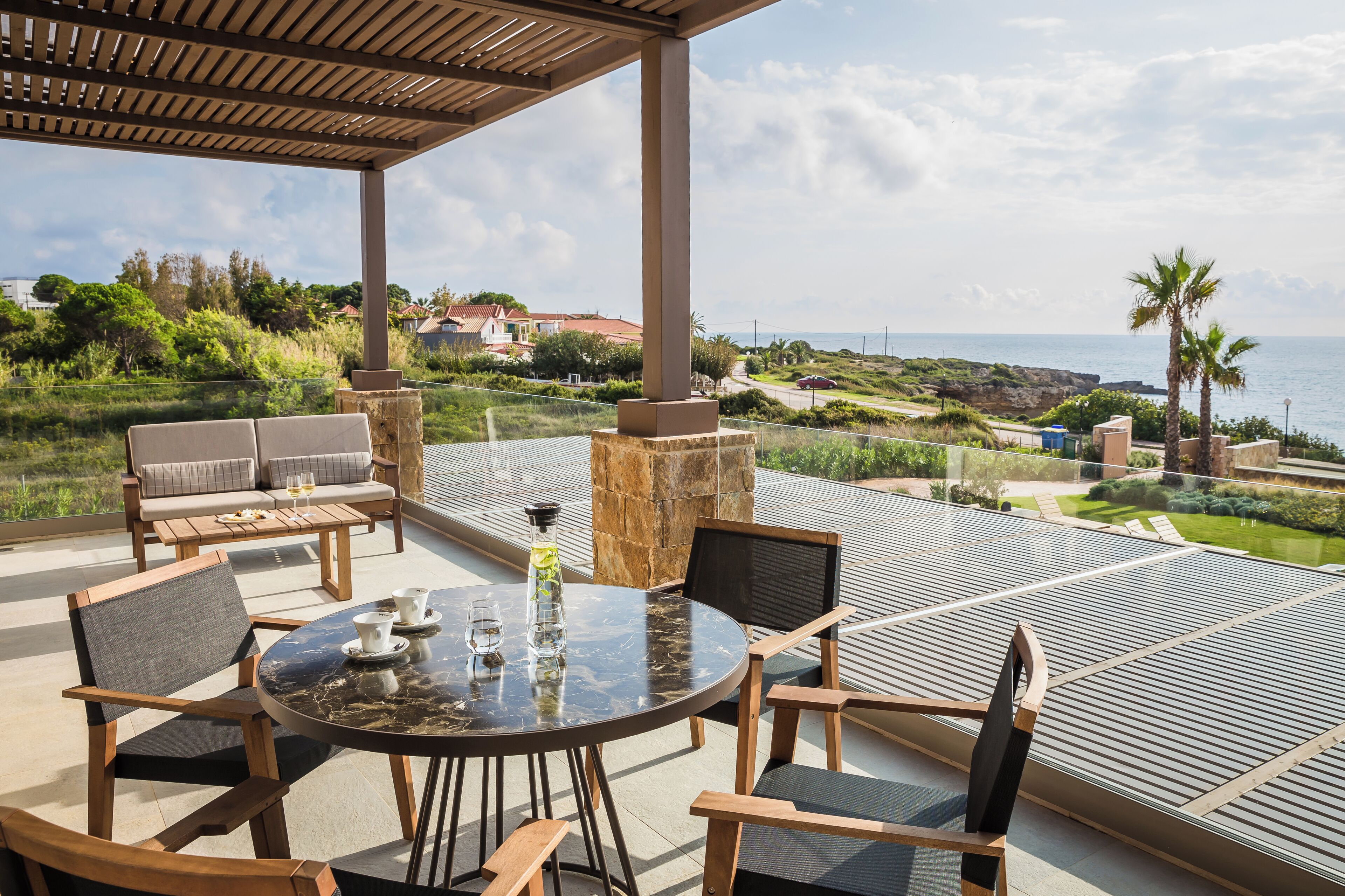 An outdoor patio with a round table, chairs, and a view of the sea and greenery.