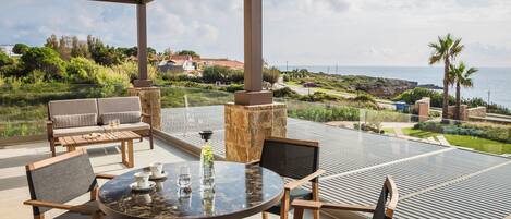 An outdoor patio with a round table, chairs, and a view of the sea and greenery.