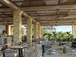 Un área de comedor al aire libre con pilares de piedra, mesas y sillas de madera, rodeada de plantas en macetas y vistas a una zona residencial.