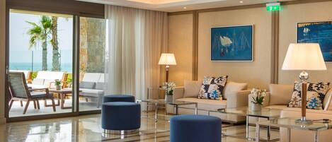 A modern hotel lobby with a glass coffee table, blue ottomans, a sofa set, and a view of the ocean through large windows.