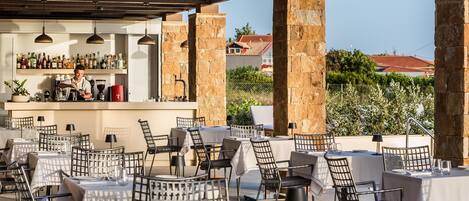 An outdoor dining area with tables and chairs, a bar with a bartender, and a view of a red-roofed building in the background.