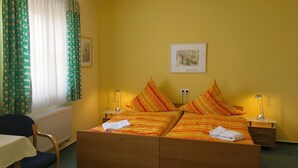 Habitación de hotel con cama de madera, dos mesitas de noche con lámparas, una silla, una mesa y una ventana con cortinas.