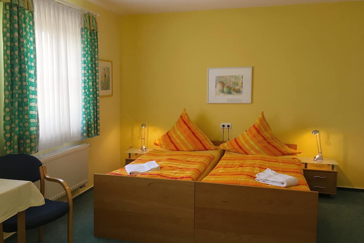 Habitación de hotel con cama de madera, dos mesitas de noche con lámparas, una silla, una mesa y una ventana con cortinas.