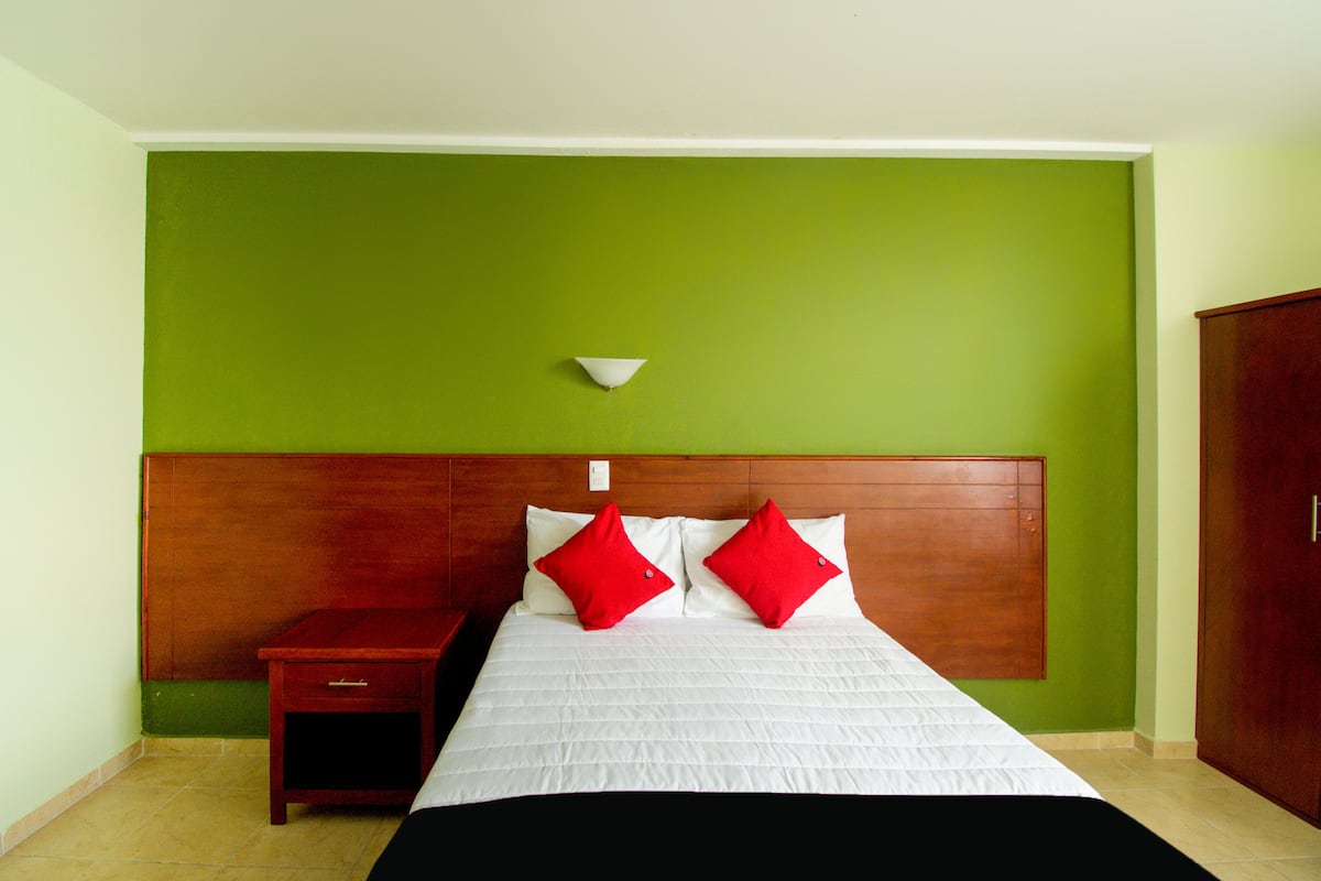 A hotel room with a green wall, a wooden headboard, a white bed with red pillows, a small wooden nightstand, and a white wall-mounted light fixture.