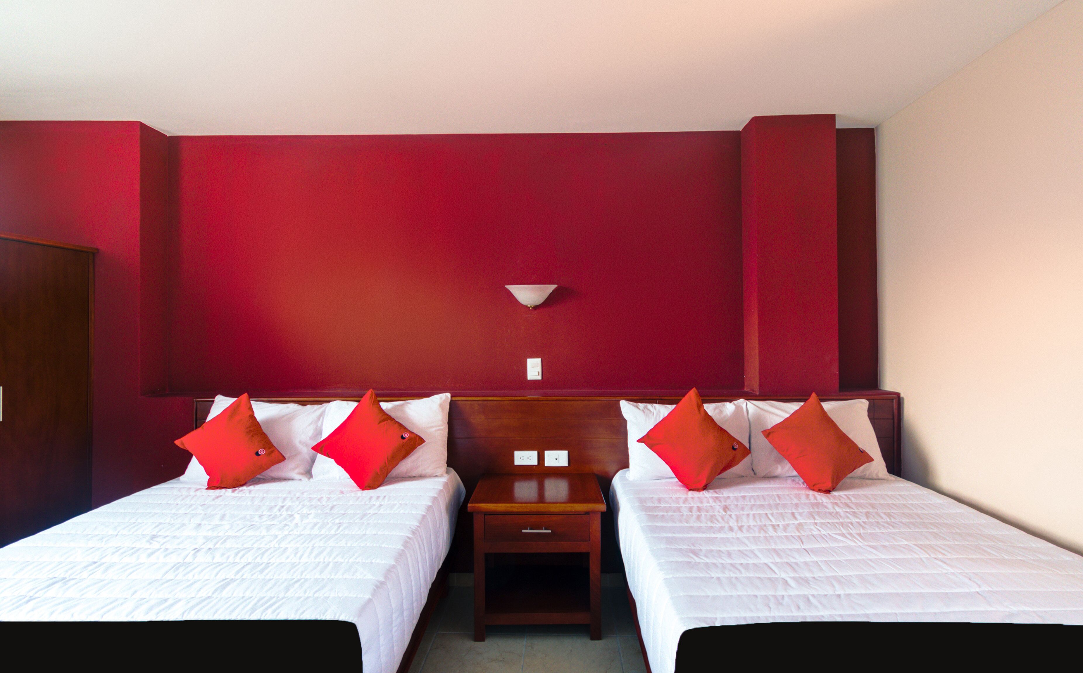 Quarto de hotel com duas camas, paredes vermelhas, mesa de cabeceira e luminária fixa na parede.