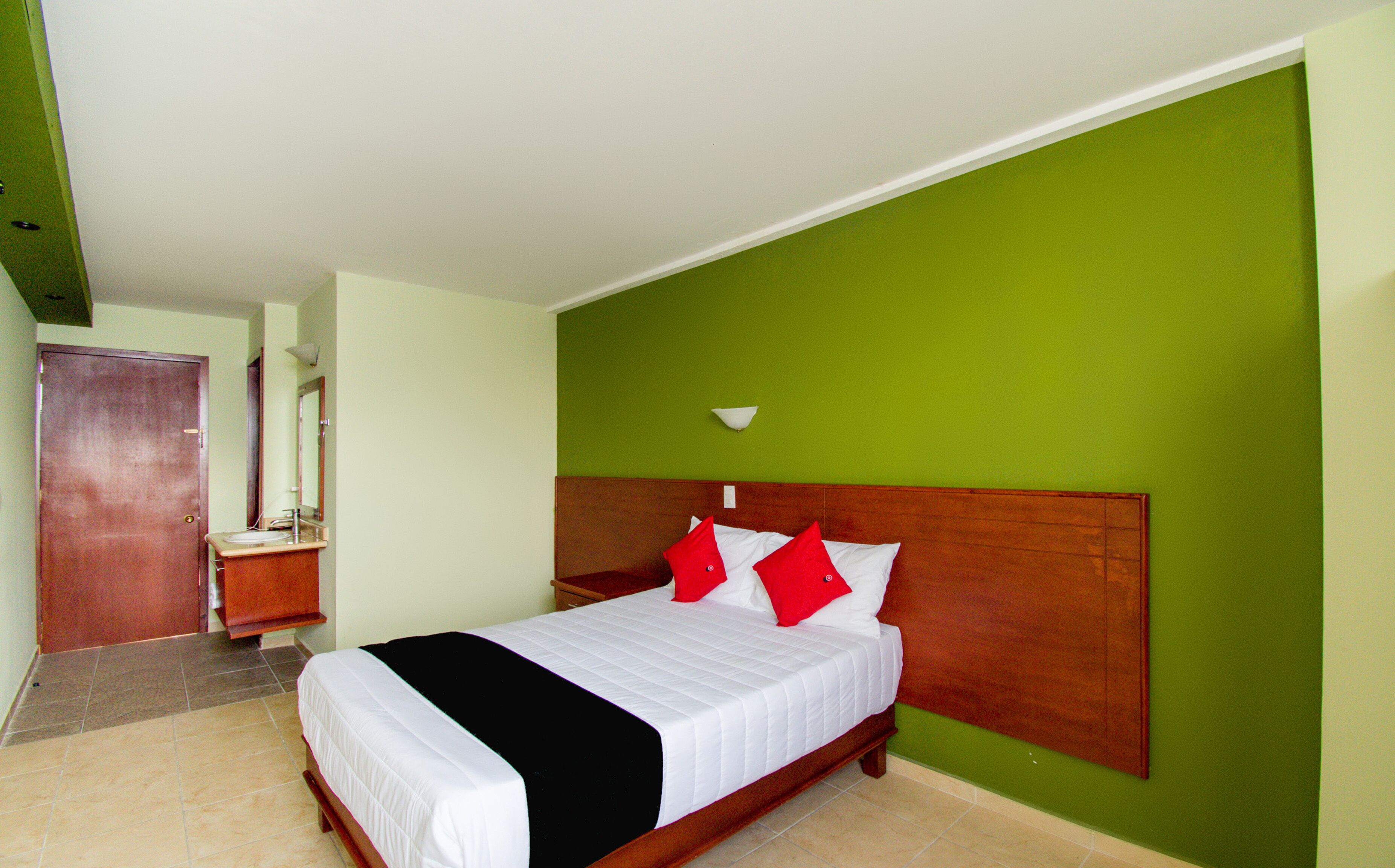 Habitación de hotel con cama, cabecera de madera, pared verde y baño con espejo y lavamanos.