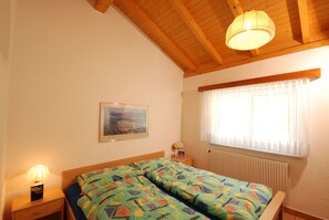 Une chambre avec un plafond en bois, un lit avec une literie à motifs, une lampe de chevet, un tableau encadré au mur et une fenêtre avec des stores.