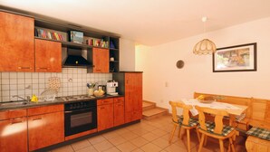 A kitchen with wooden cabinets, a stove, and a dining area with a table and chairs. - Casa Esmeralda SERENITA - 3-room apartment, approx. 65 m², with garden seating area (Gambarogno)