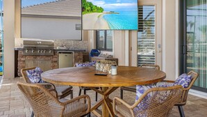 An outdoor patio with a round wooden table, wicker chairs, a grill, and a TV screen displaying a beach scene. - Wonderful New 3 Bedroom Villa , with direct Gluf Acces for your holidays (CAPE CORAL)