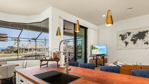 A modern kitchen with a wooden countertop and a sink, a dining area with blue chairs, and a large window offering a view of palm trees and a house. - Wonderful New 3 Bedroom Villa , with direct Gluf Acces for your holidays (CAPE CORAL)