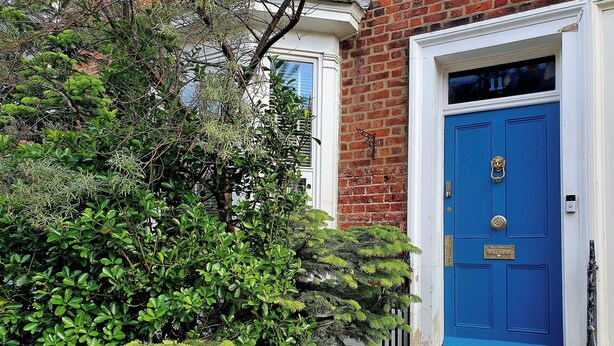 Une porte bleue ornée d’un heurtoir en laiton, un mur de briques et un petit jardin planté de différentes espèces.