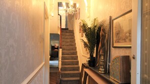 Un couloir avec un escalier, un lustre, des œuvres d’art encadrées et une plante en pot.