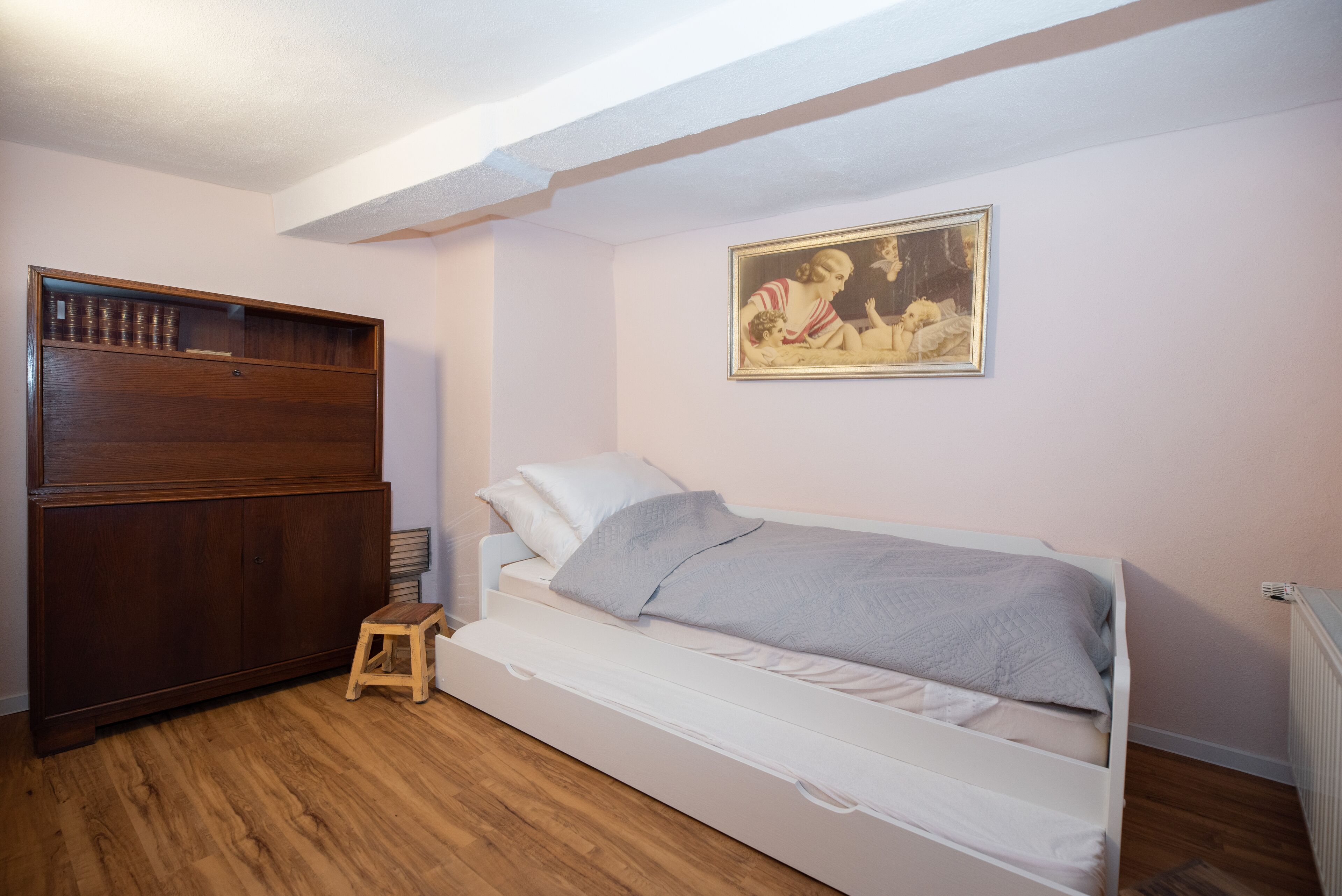 Un dormitori amb un llit, un armari de fusta, un petit tamboret i un quadre al mur.