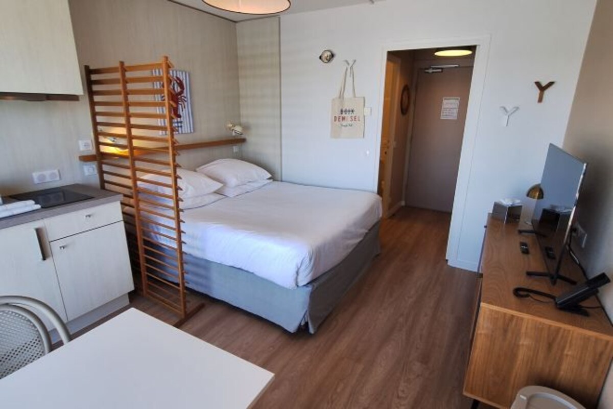 A compact hotel room with a bed, a desk with a computer, a chair, a small kitchenette, and a wooden ladder.