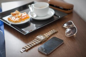 A tray with a cup of coffee, a plate of food, a watch, a smartphone, and a pair of sunglasses on a wooden table. - Hotel Bologna (Misano Adriatico)